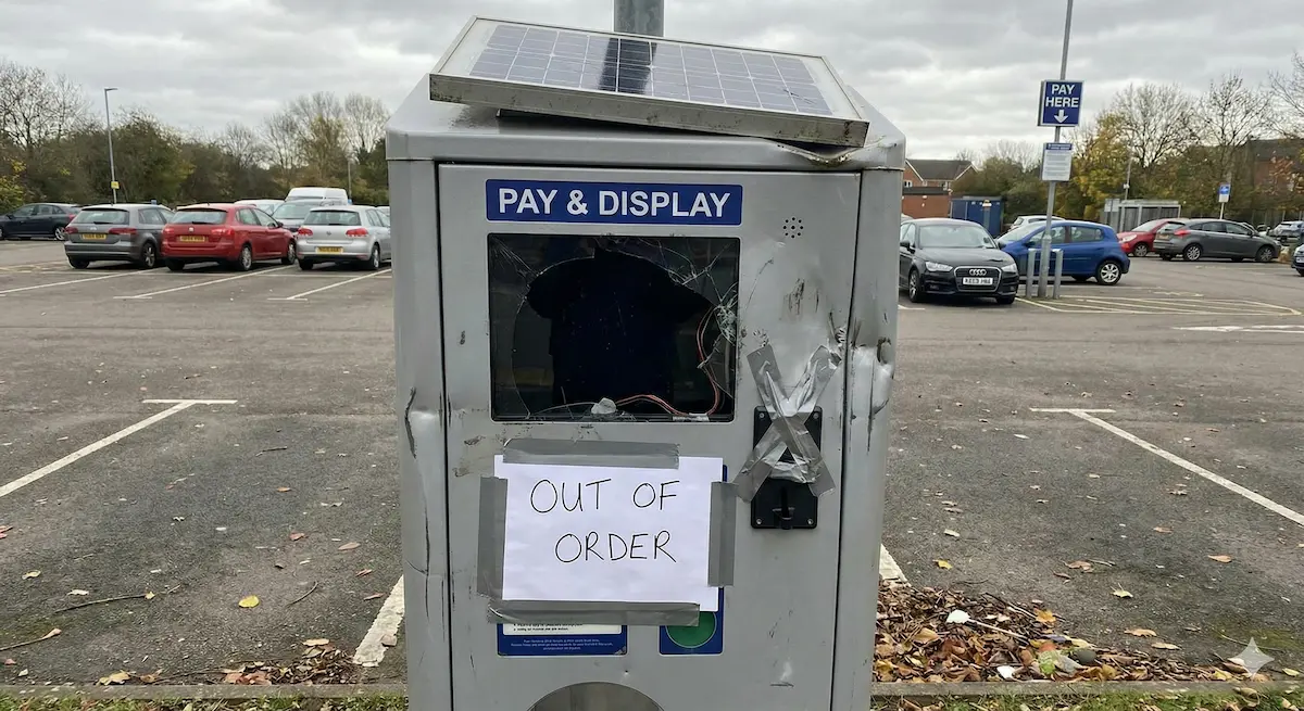 Damaged pay and display parking meter with cracked screen and a taped sign saying 'OUT OF ORDER' in an empty parking lot.