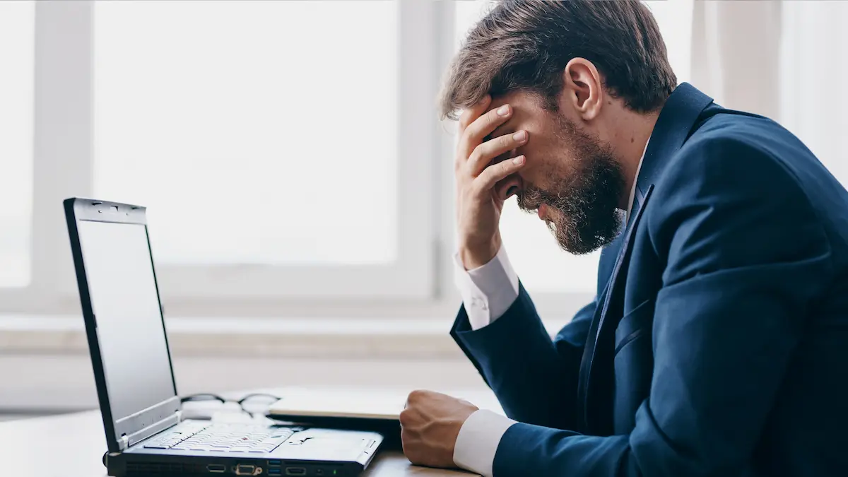 Frustrated man in a suit sitting at a desk with his hand on his forehead in front of a laptop.