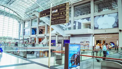 Interior view of a modern shopping mall with high ceilings, escalators, and directional signs for shops and services.