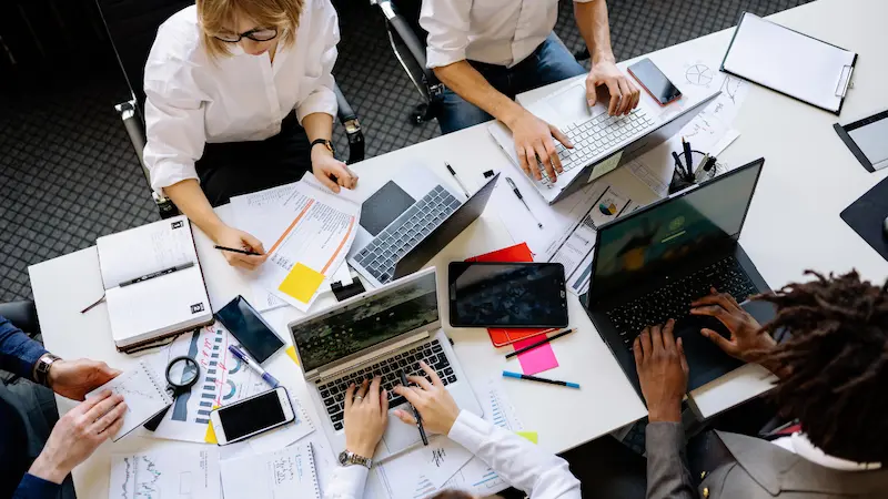 Overhead view of diverse team collaborating at office desk with laptops, papers, and tablet.