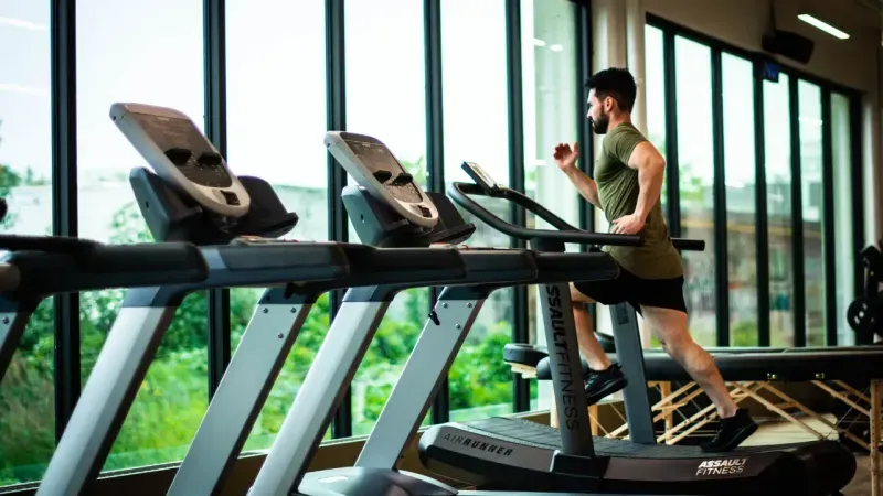 Man running on a treadmill in a gym with large windows showing green outdoor scenery.