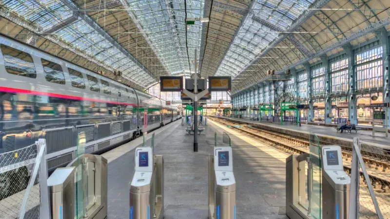 Modern train station platform with transparent ticket barriers and a high-speed train on the left under a large glass roof.