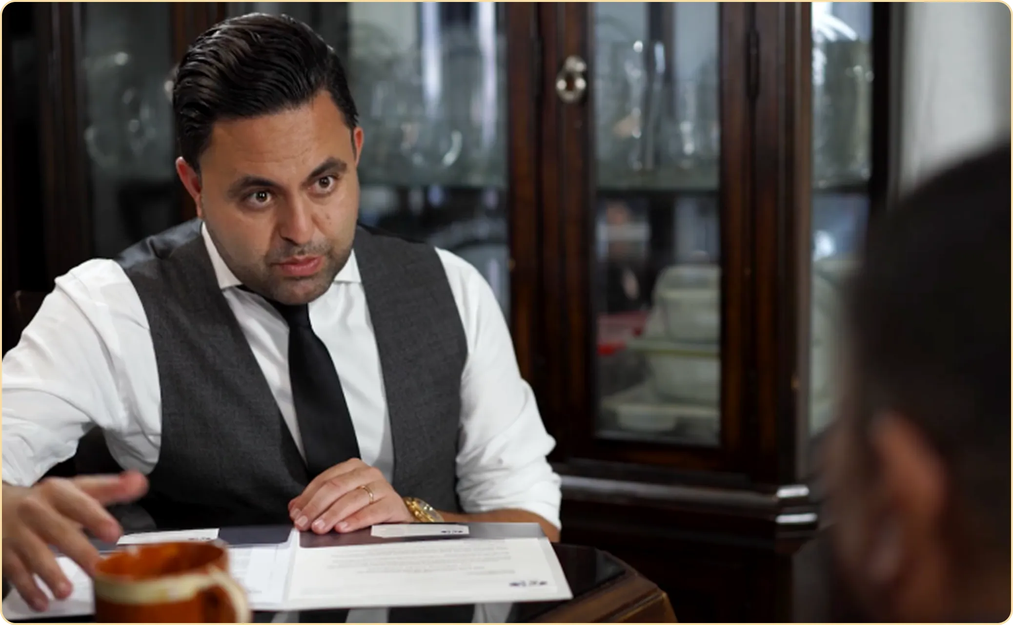 Man in white shirt, black tie, and gray vest sitting at a table, discussing documents with another person.