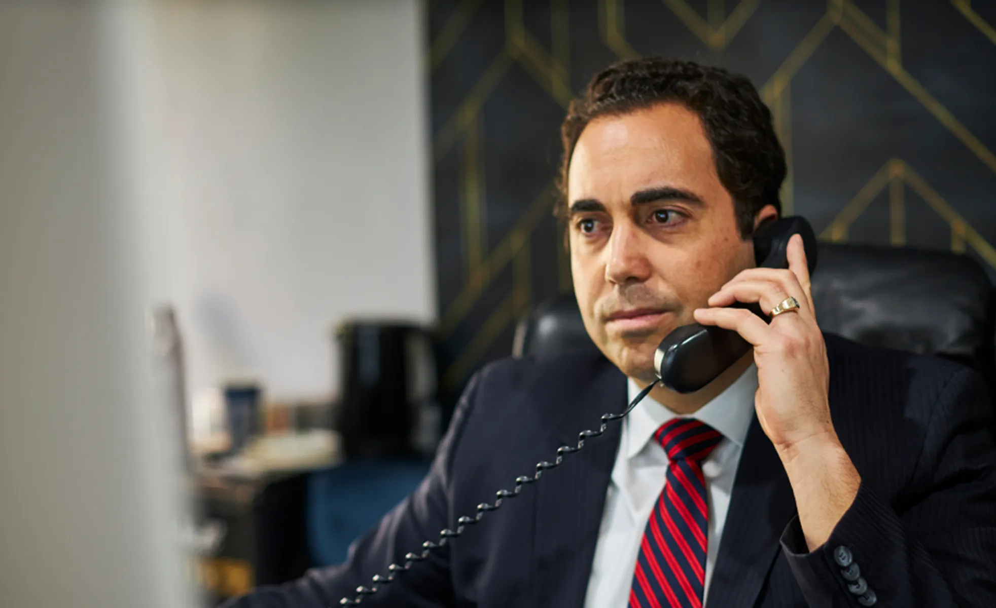 Man in a suit with a red and blue striped tie talking on a corded telephone in an office.