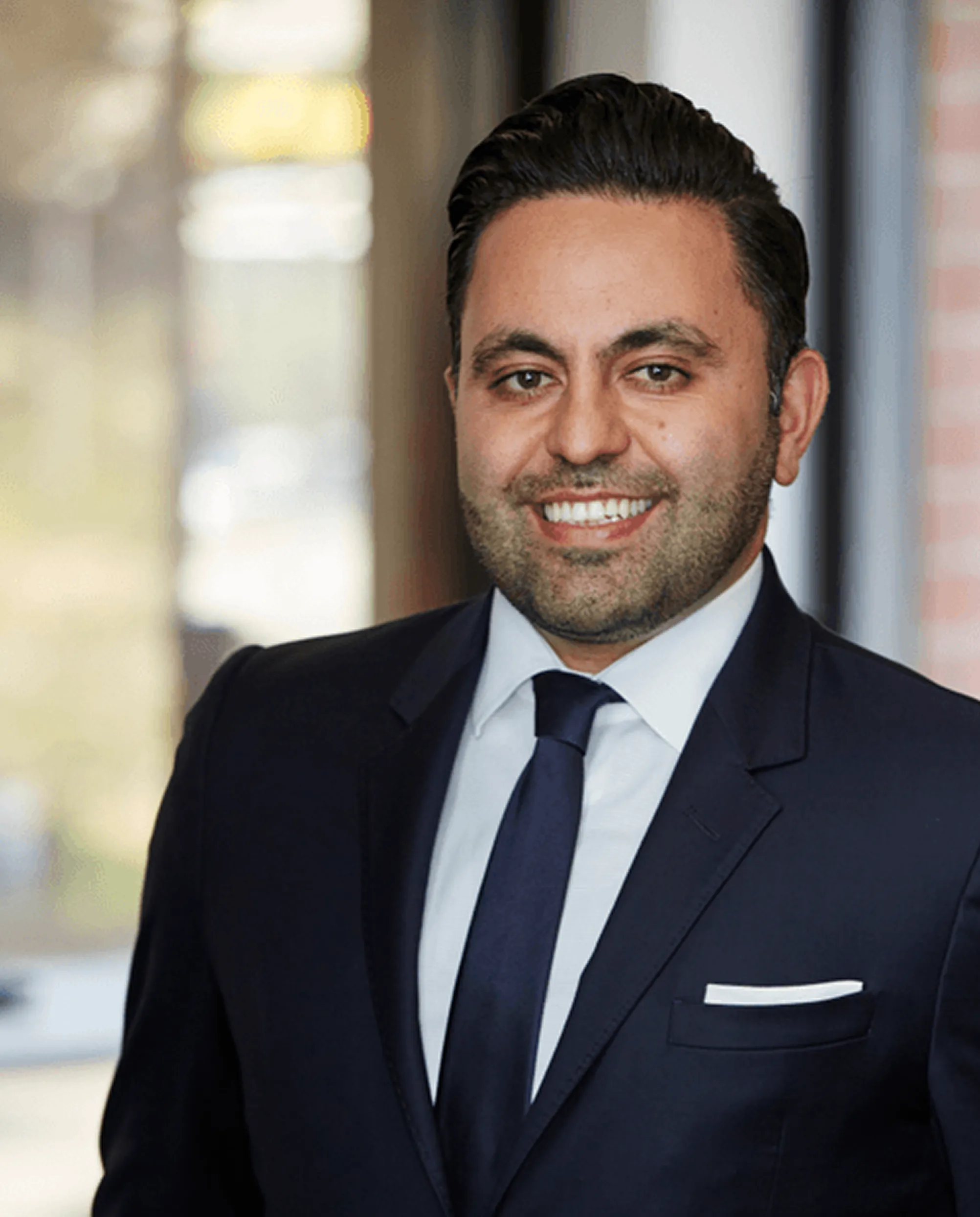 Smiling man with dark hair wearing a navy suit, white shirt, and navy tie indoors.