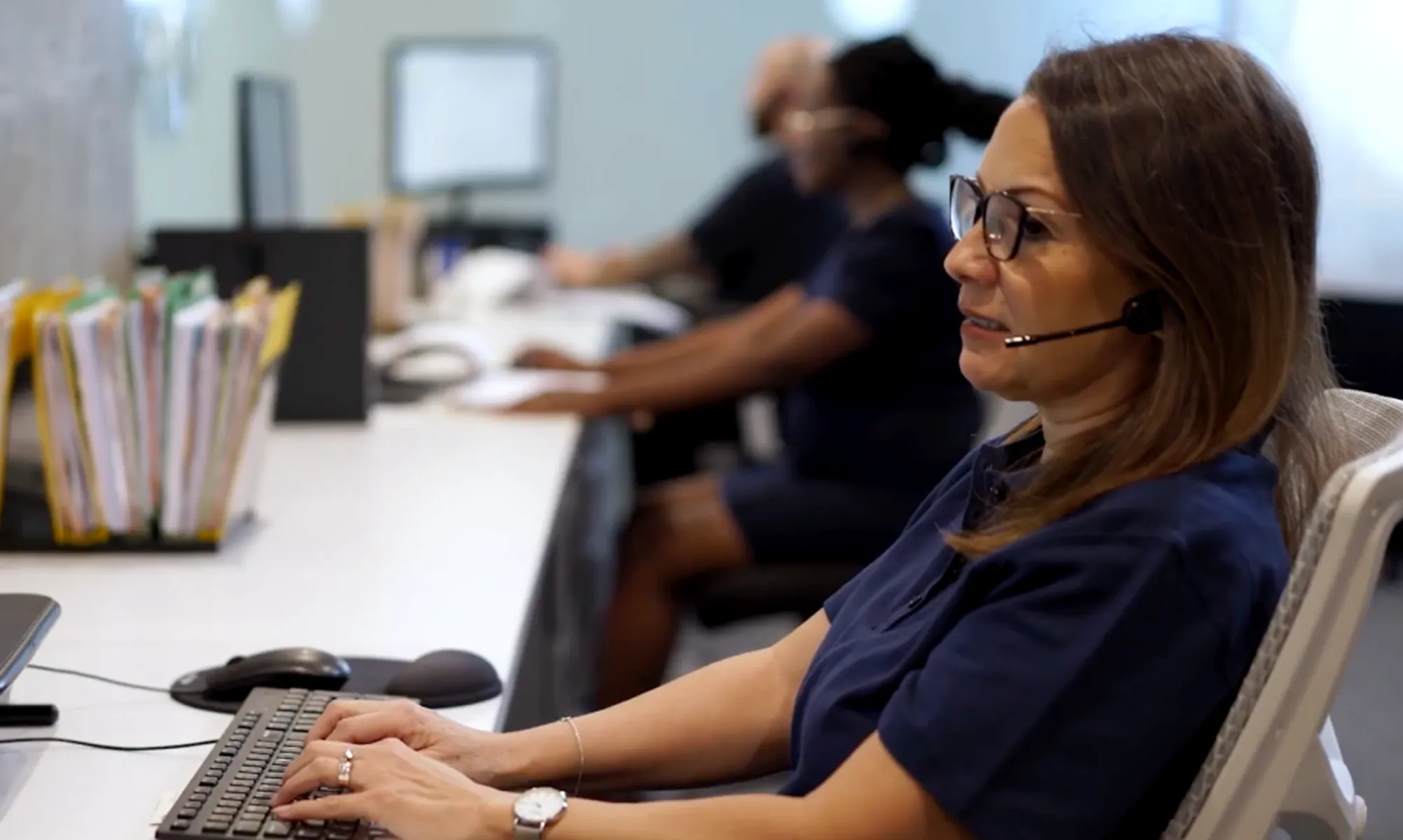 Woman wearing glasses and headset typing on a keyboard in an office with colleagues at desks.