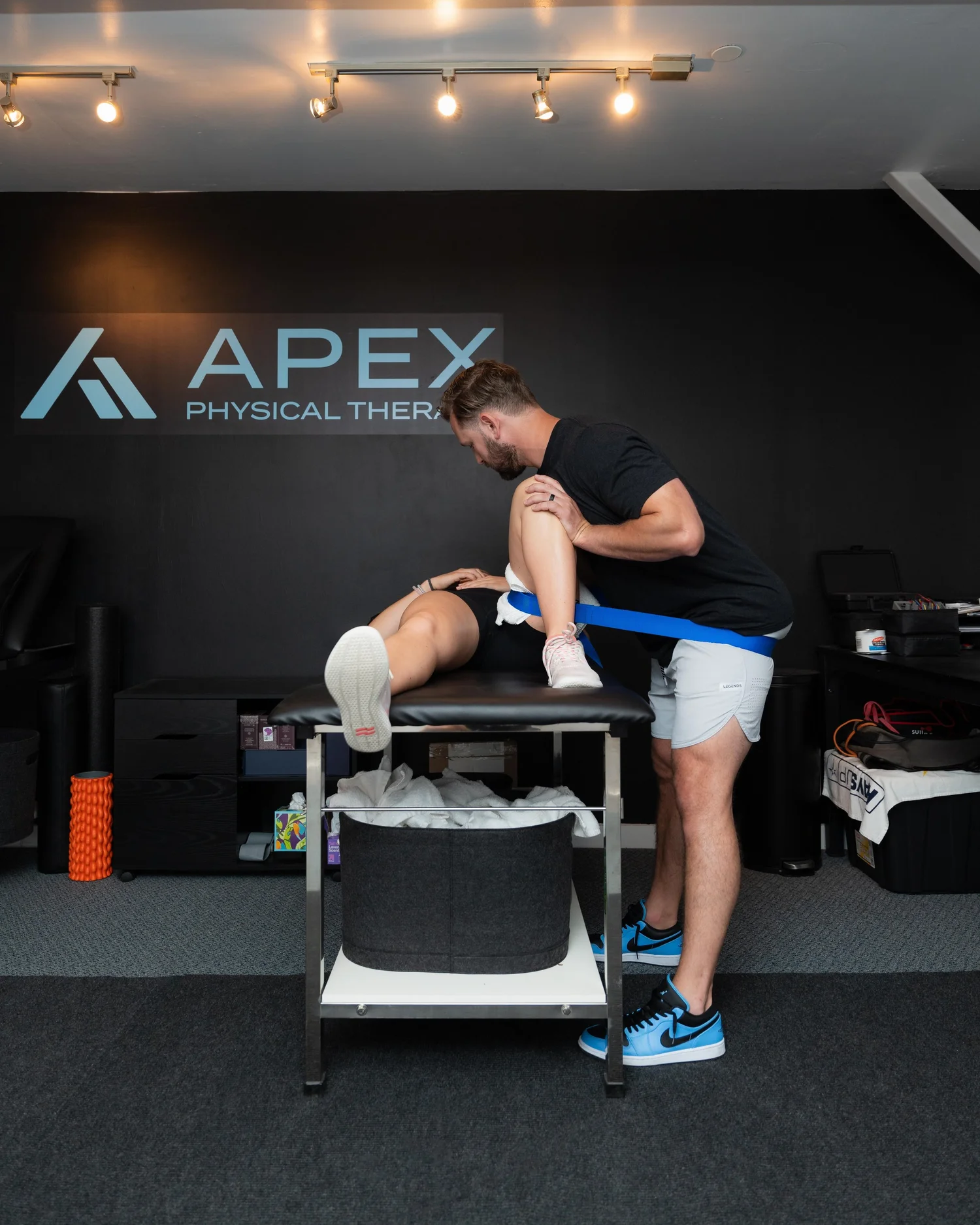 Physical therapist assisting a patient with leg exercise using a resistance band in a treatment room with Apex Physical Therapy logo on the wall.