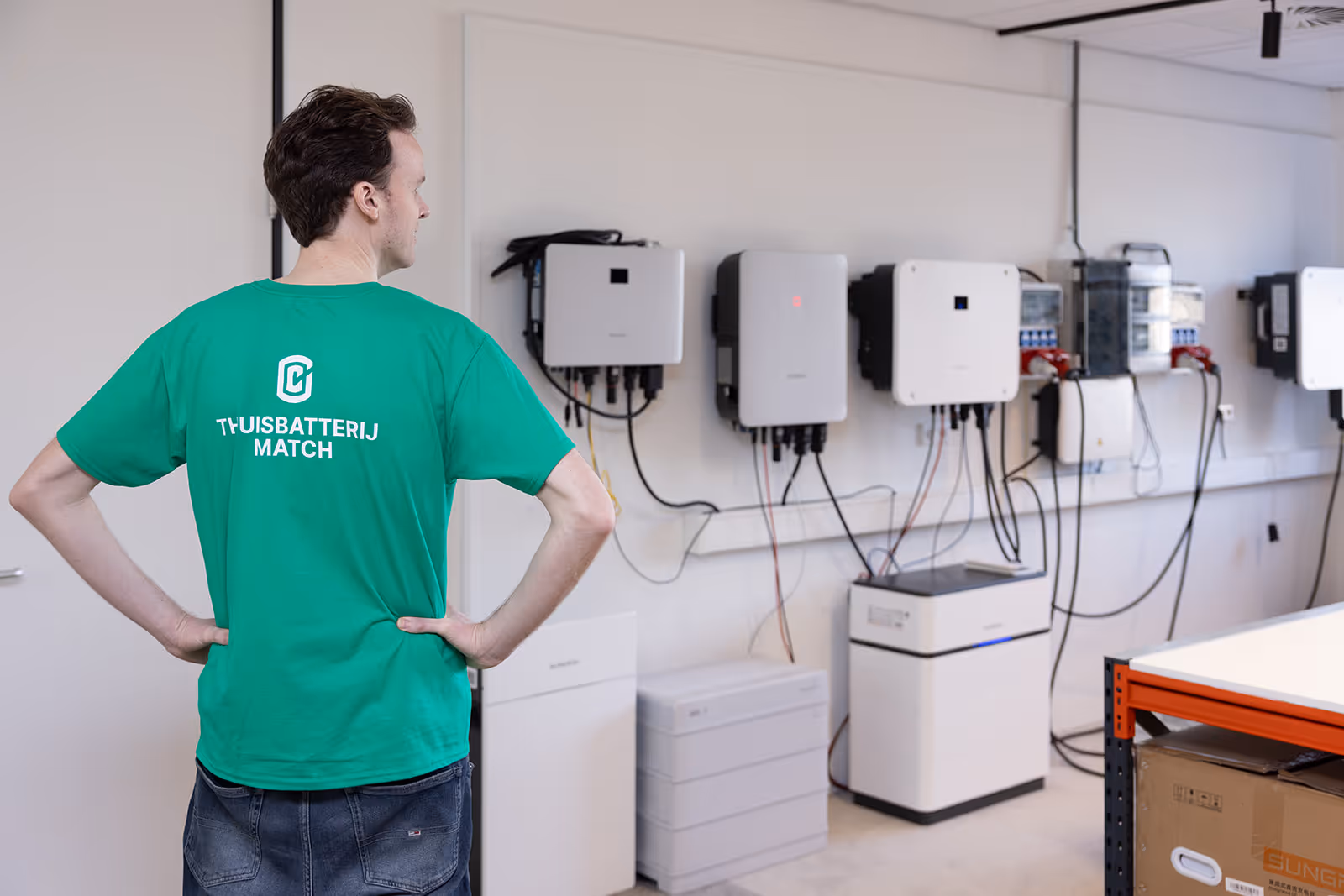 Man met groen T-shirt met tekst 'THUISBATTERIJMATCH' kijkt naar een muur met meerdere thuisbatterijen.