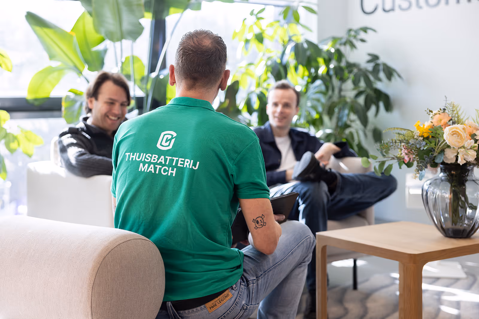 Man met groen shirt met tekst 'ThuisbatterijMatch' praat met twee mannen in een lichte kamer met veel planten.