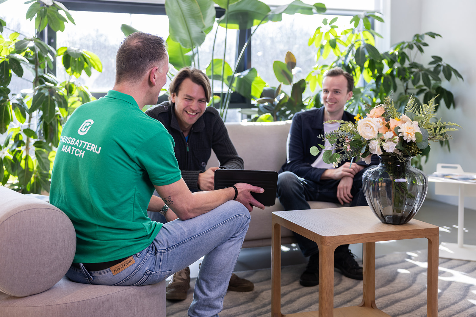 Drie mannen zitten in een lichte kamer met planten en praten ontspannen op banken, waarbij een man in een groen shirt met het logo ‘ThuisbatterijMatch’ een tablet vasthoudt.