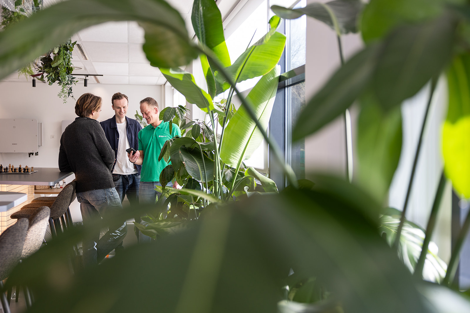 Drie mannen praten en lachen samen in een kantoorruimte met veel groene planten bij een raam.