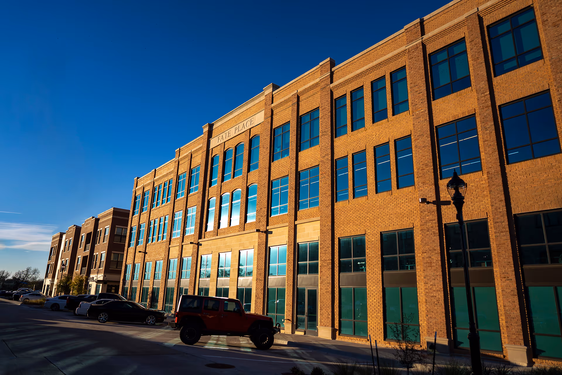 Three-story brick building named Fate Place with large windows reflecting a clear blue sky, parked cars and a street lamp in front.