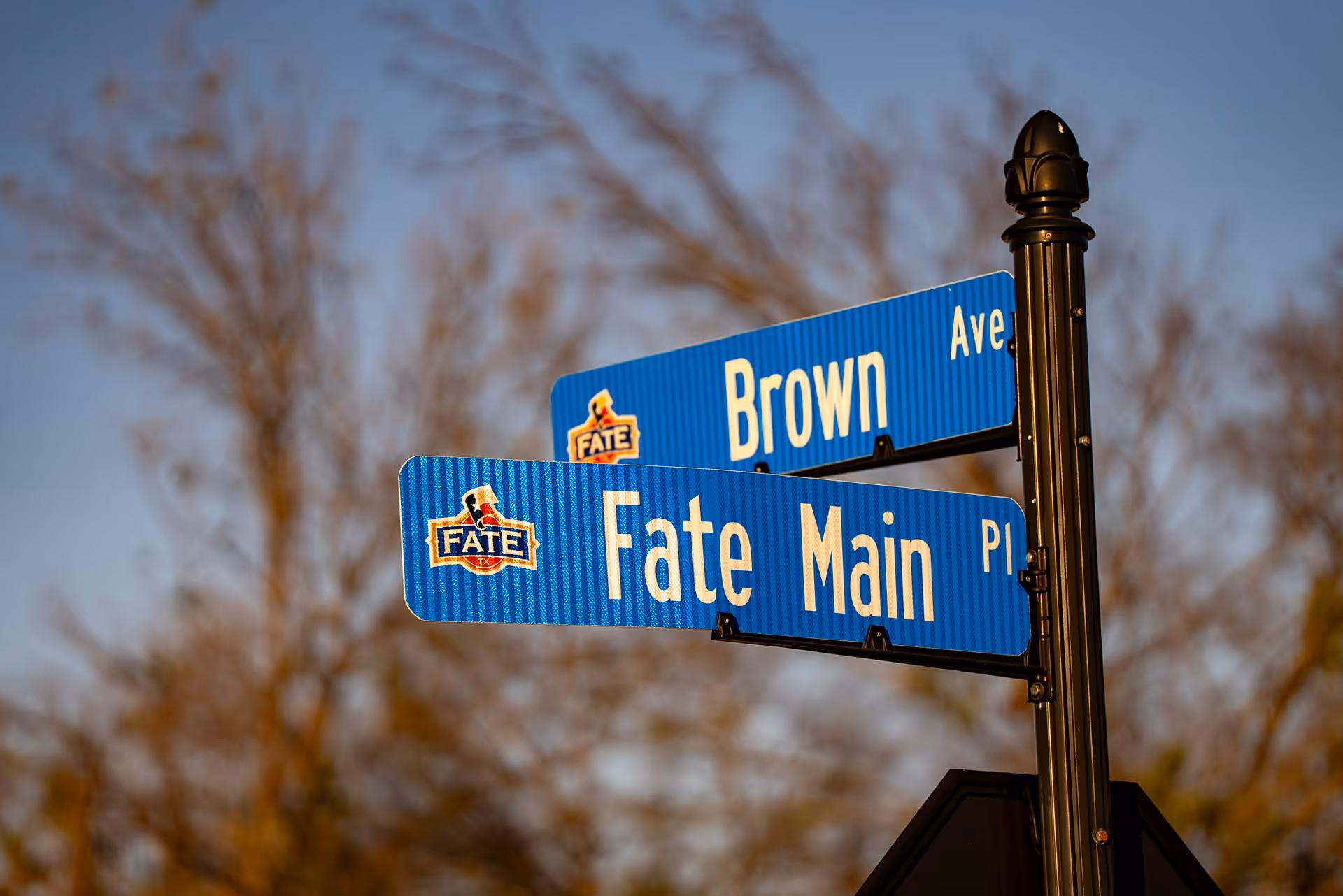 Blue street signs at an intersection showing Brown Ave and Fate Main Pl with a Fate Texas logo on each sign.