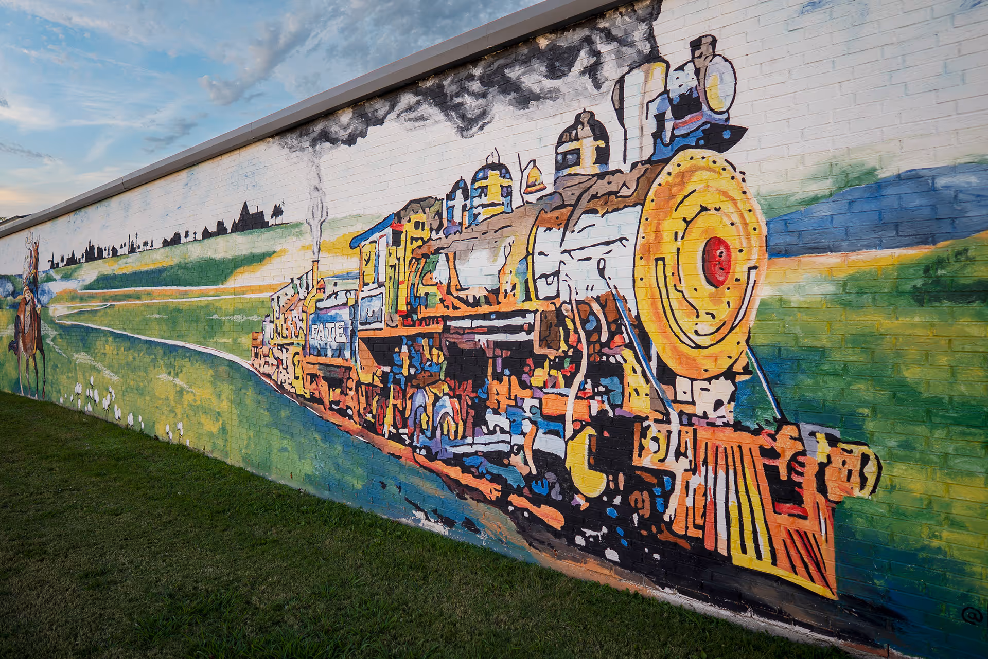 Colorful mural of a steam locomotive train with smoke on a brick wall surrounded by green grass and a rural landscape.
