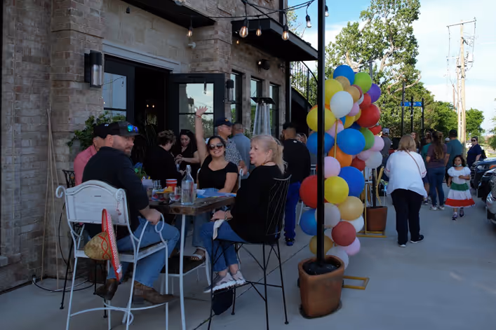 People gathered outside a brick building, sitting and standing near tables decorated with colorful balloons during a sunny day.