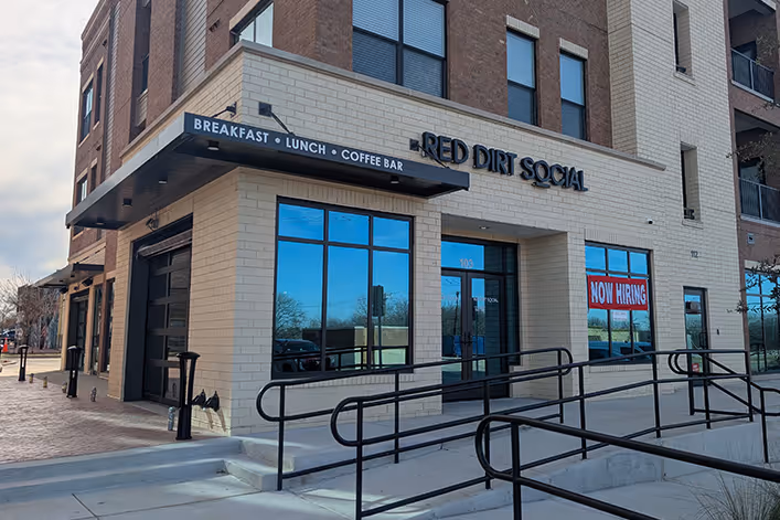 Corner view of a brick building with signage for Red Dirt Social and a 'Now Hiring' sign in the window.