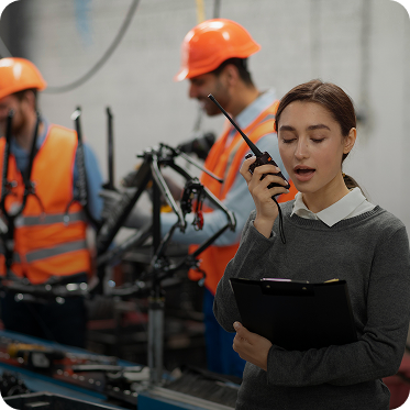 Woman speaking into a walkie-talkie holding a clipboard in a factory setting with workers in orange safety vests and helmets.