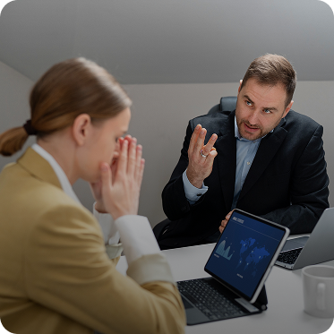 Two business professionals in discussion, one appearing thoughtful and the other explaining with a hand gesture, with a tablet displaying a world map on the table.
