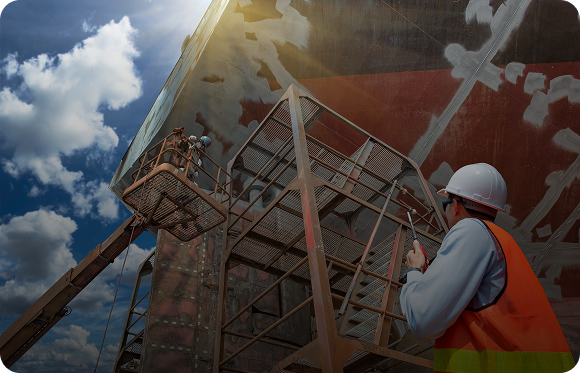 Construction worker in a hard hat and orange safety vest operating equipment near a large industrial structure under a partly cloudy sky.