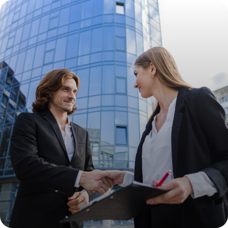 Businesswoman and businessman shaking hands outside a glass office building, holding a clipboard and pen.