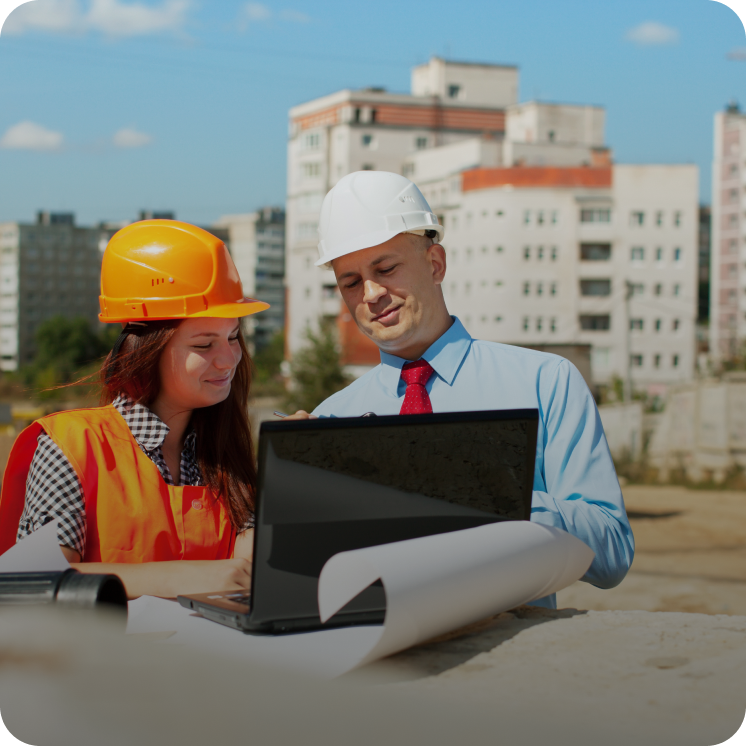 Male and female construction workers wearing helmets reviewing documents and laptop on a construction site with buildings in the background.