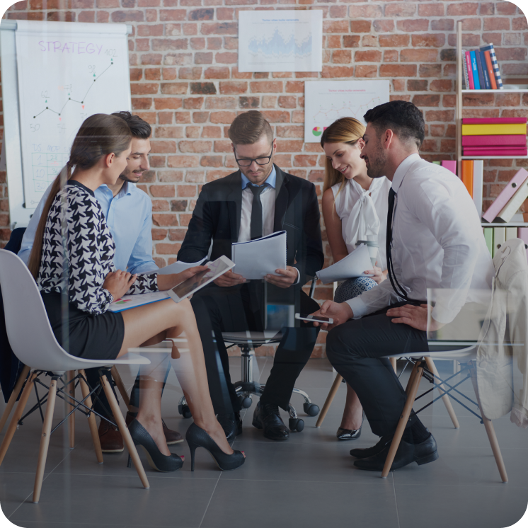 Five young professionals engaged in a collaborative meeting in an office with a brick wall and charts in the background.