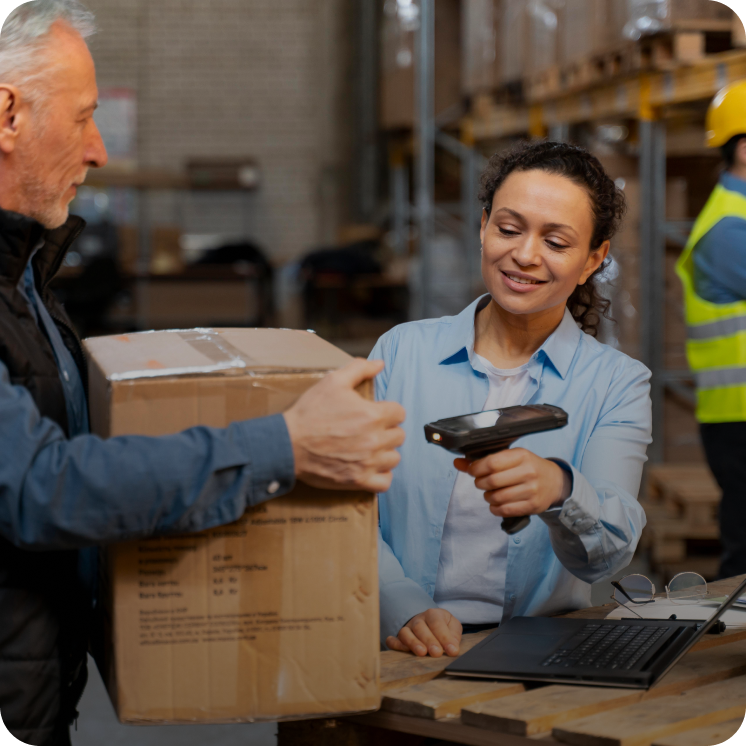 A woman scans a cardboard box with a handheld barcode scanner held by a man in a warehouse setting.