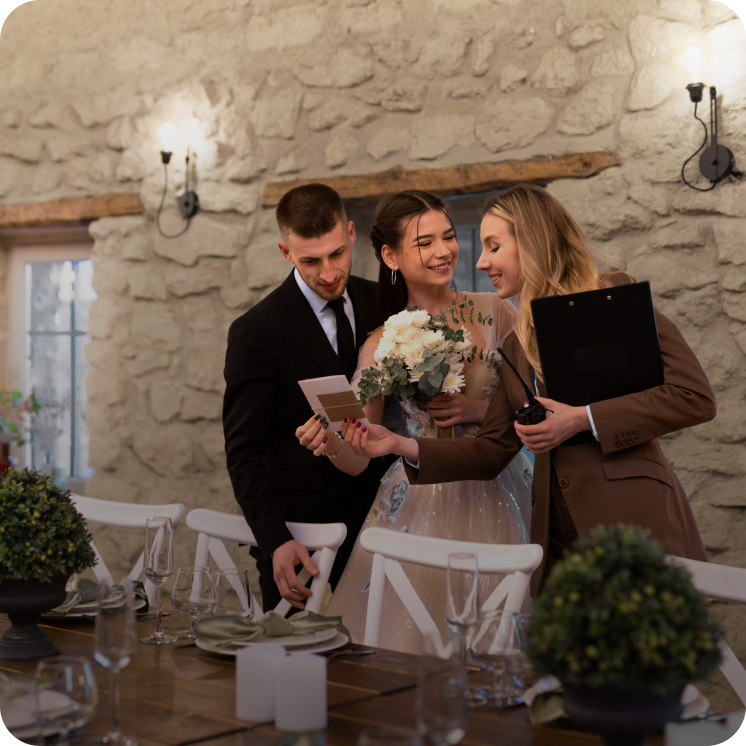 Bride and groom consulting with a wedding planner in a stone-walled venue, with the bride holding a bouquet and the planner showing a card.