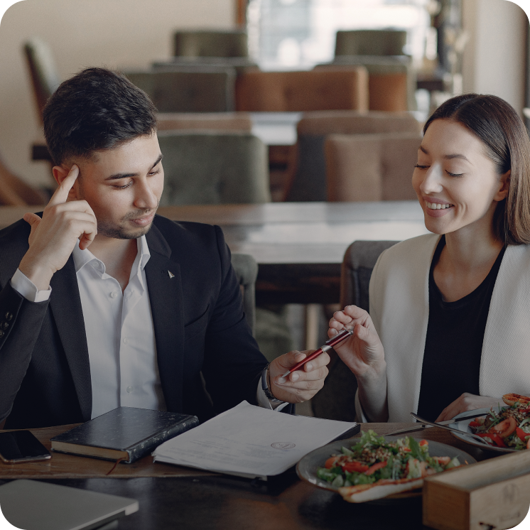 Man and woman sitting at a table with documents and salads, exchanging a pen during a discussion in a restaurant setting.