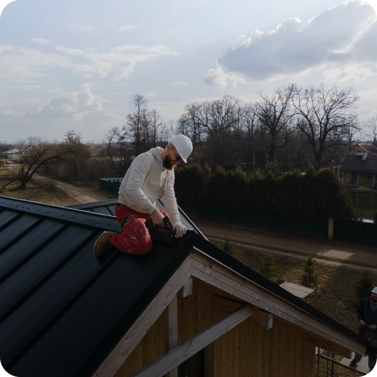 Construction worker wearing a white hard hat and gloves, kneeling on a black metal roof while using a cordless drill on a wooden house.