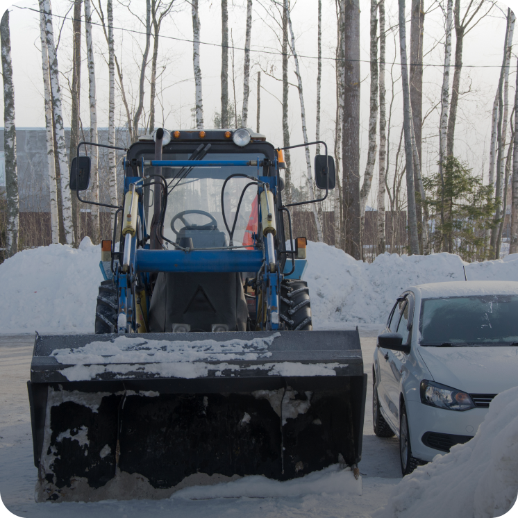 Blue snow plow vehicle parked next to a white car with snow-covered ground and bare trees in the background.