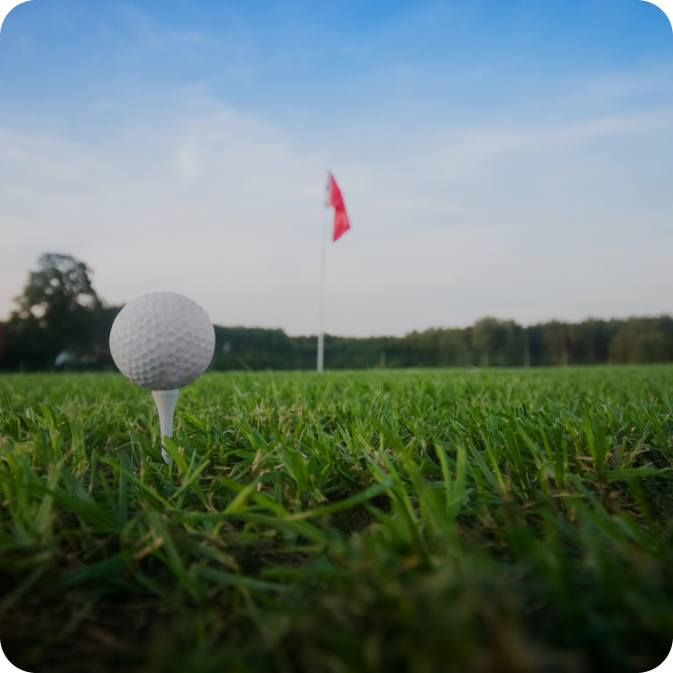 Close-up of a golf ball on a tee with a red flag in the distance on a golf course under a blue sky.