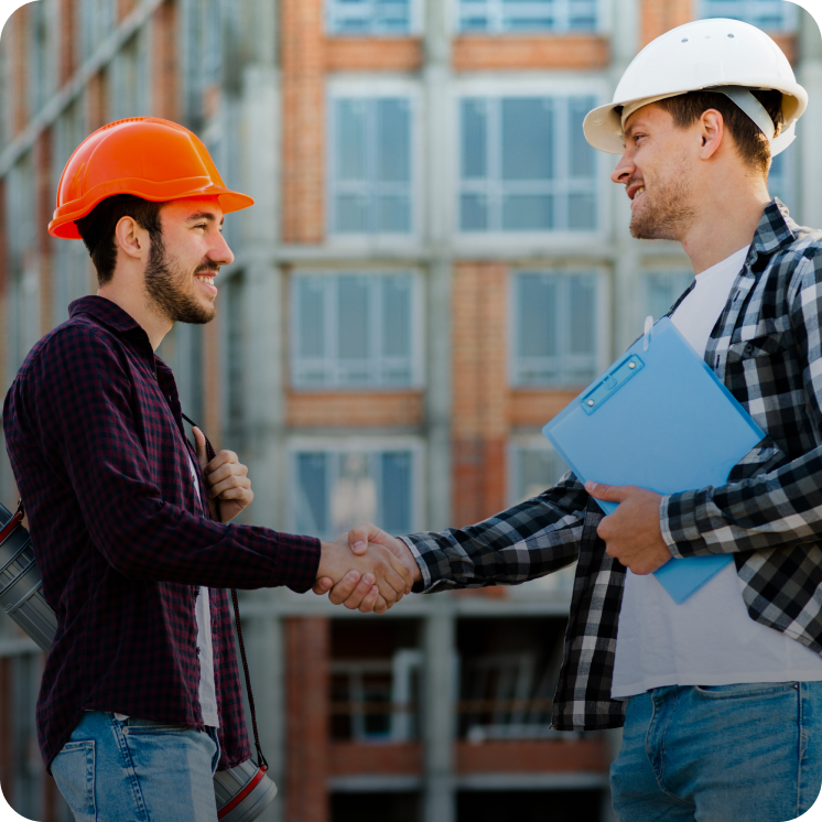 Two construction workers wearing hard hats shaking hands in front of a building under construction.
