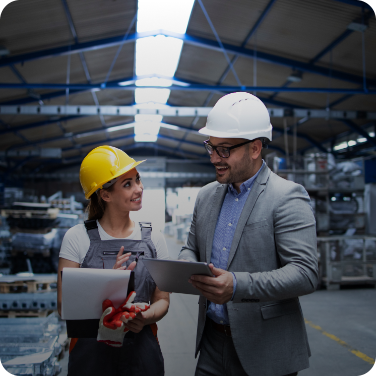 Two workers in a warehouse wearing hard hats, one holding a clipboard and the other a tablet, discussing work.