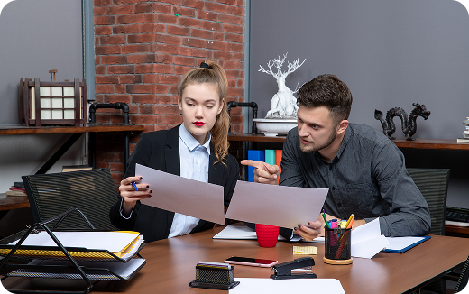 Two colleagues sitting at a table reviewing documents, with office supplies and a laptop on the table.