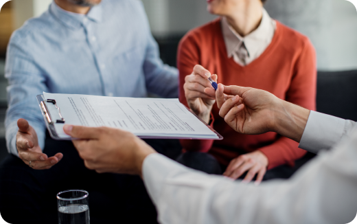 Person handing a clipboard with documents to a couple, while another hand offers a pen for signing.