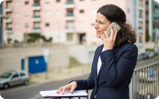 Businesswoman in a navy blazer talking on a smartphone while holding documents outdoors near a railing.