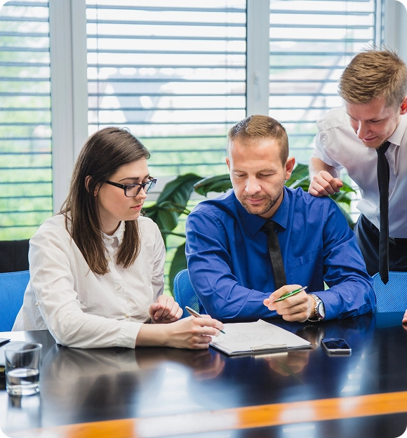 Three business professionals in formal attire discussing and reviewing documents at a conference table.