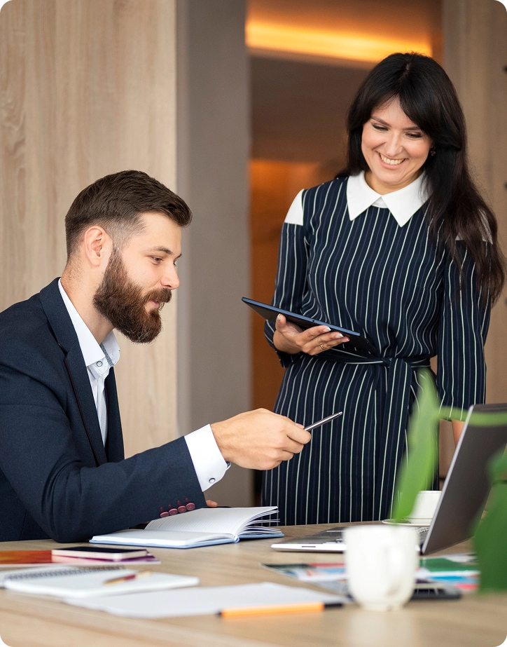 Man in suit sitting at a desk pointing at a laptop screen with a pen while a woman in a striped dress stands beside him holding a tablet and smiling.