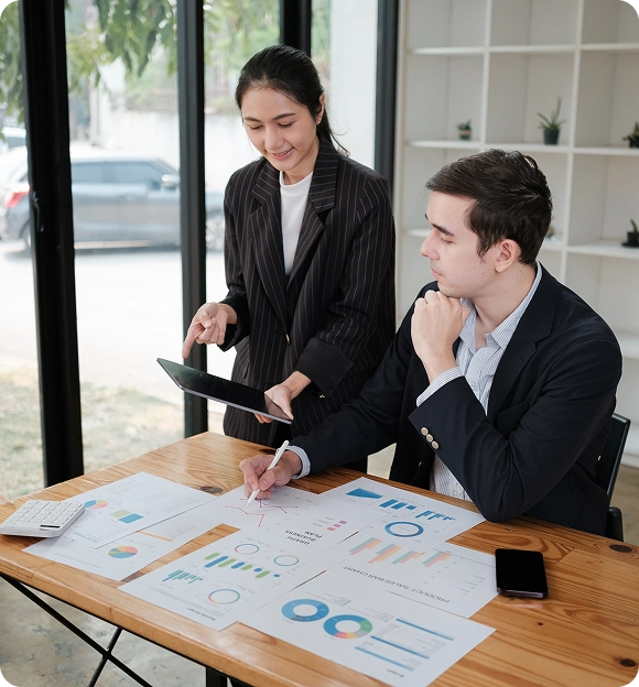 Two business professionals analyzing charts and graphs at a wooden table, one seated and writing while the other stands holding a tablet.