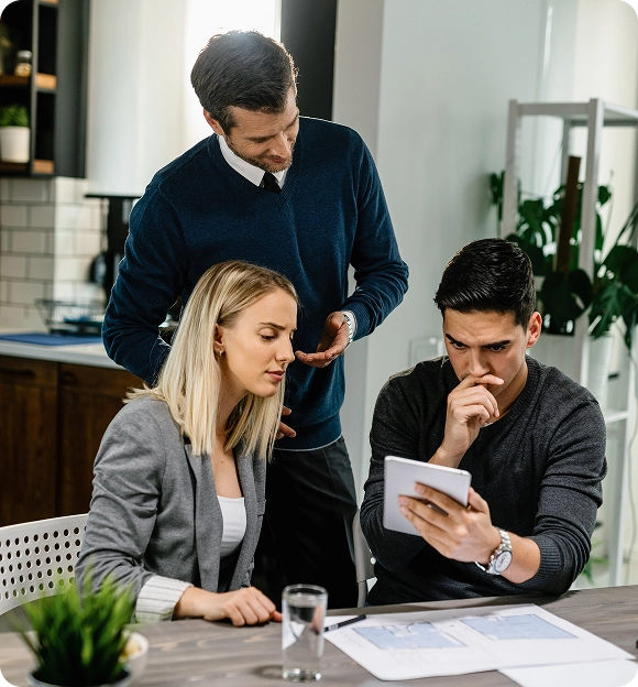 Three colleagues reviewing information on a digital tablet at a table with blueprints and a glass of water.