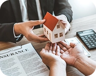 Two people exchanging a small model house over an insurance document and calculator on a desk.