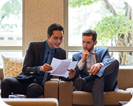 Two men in suits sitting on a couch reviewing documents together in a bright room with large windows and greenery outside.