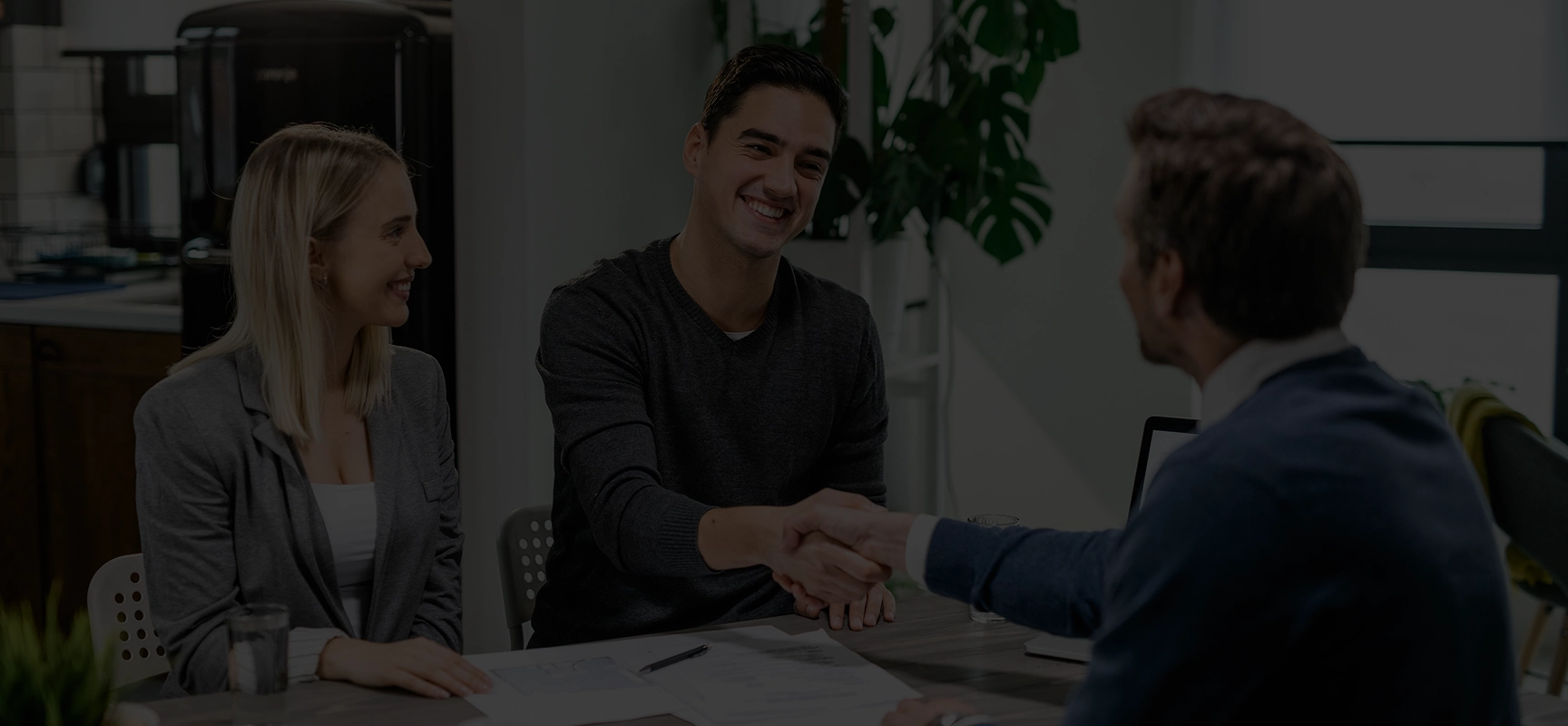 A man and woman sitting at a desk smiling and shaking hands with a professional in an office setting.