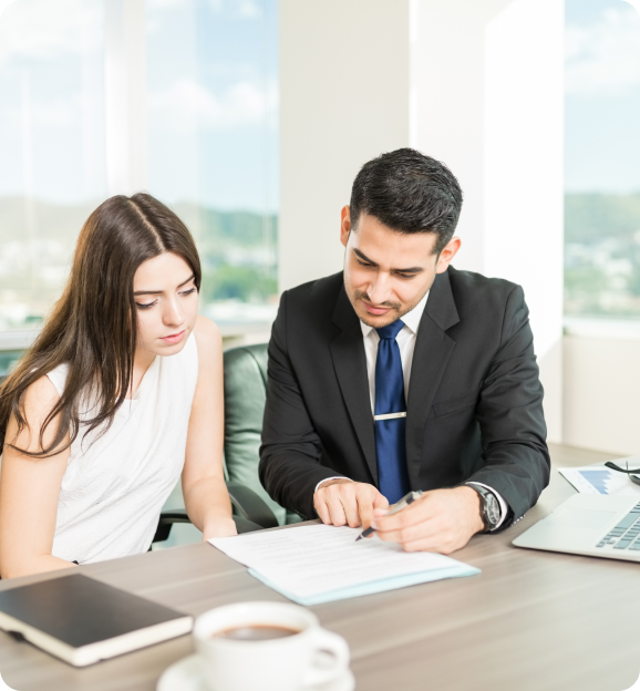 A businessman in a suit explains a document to a woman in a white sleeveless top at a desk with a laptop and coffee cup.