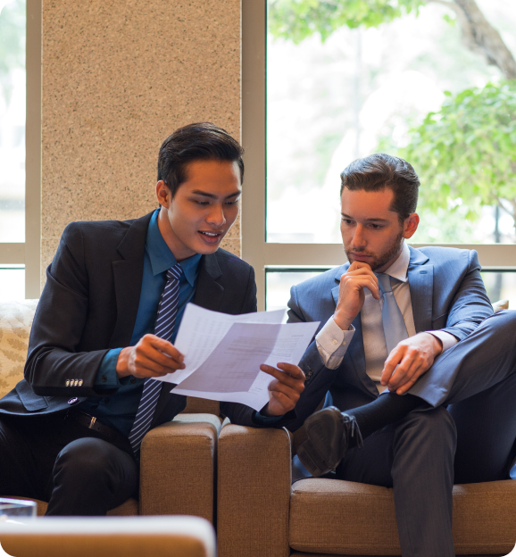 Two businessmen in suits sitting on a couch reviewing and discussing documents together.