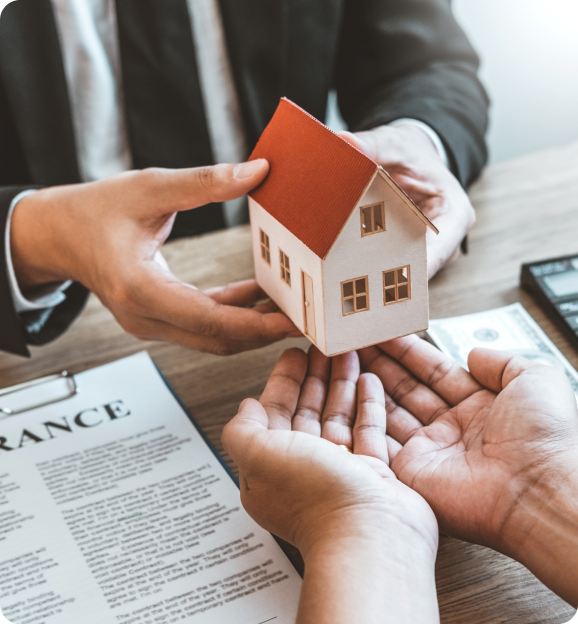 Person in suit handing a miniature house model to another person over a table with an insurance document.