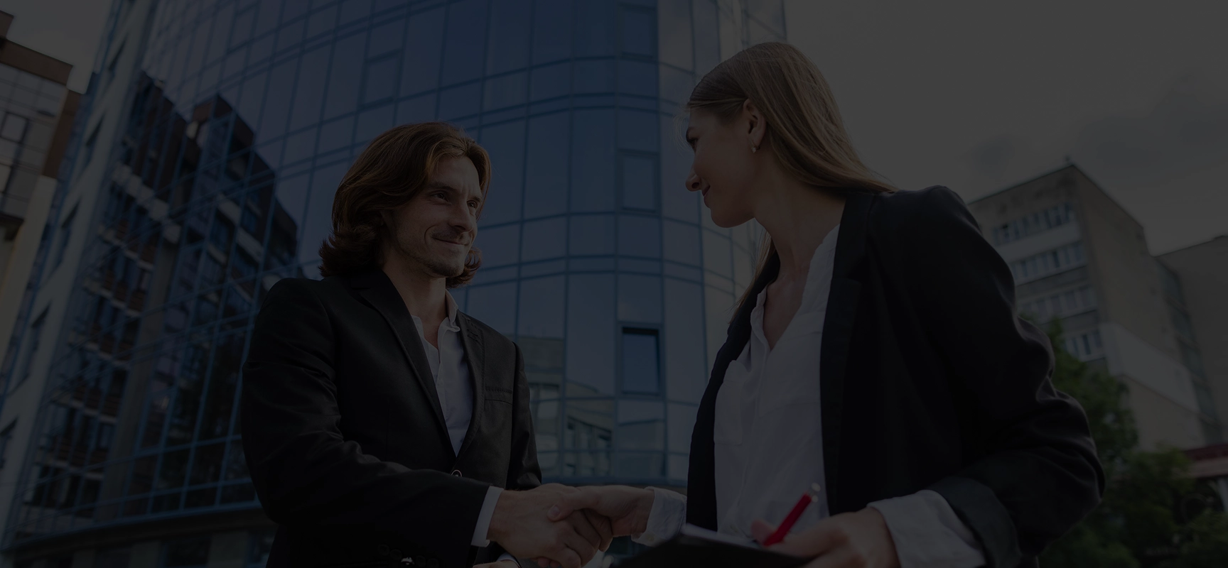 Two business professionals shaking hands outdoors in front of a modern glass office building.