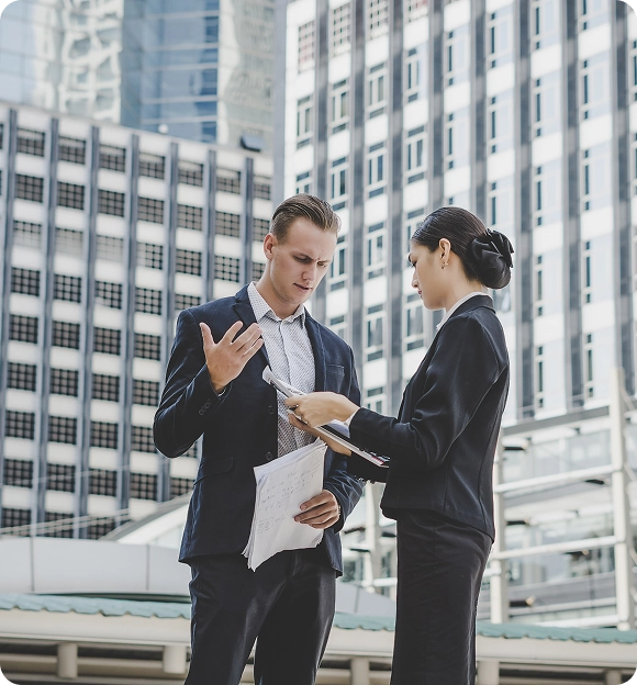 Two business professionals discussing documents outside with modern office buildings in the background.