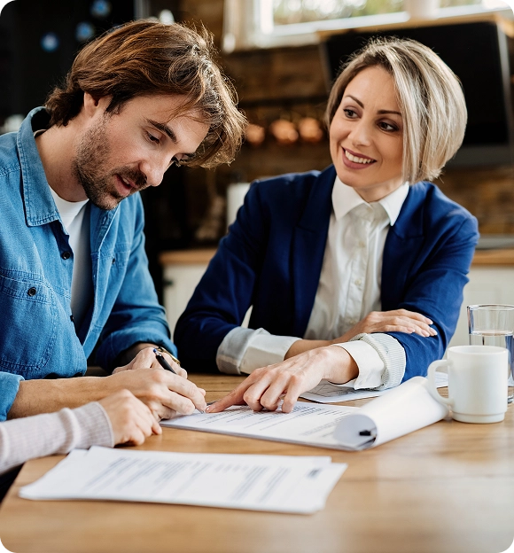 Smiling woman in a blue blazer assisting a man with signing documents at a wooden table in a cozy kitchen setting.